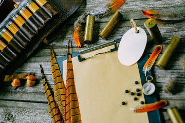 Closeup feather and cartridges on clipboard with vintage paper and oval label for information on old wooden background. Hunting equipment on vintage board