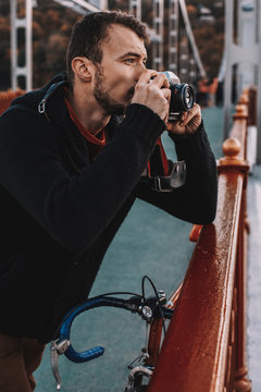 Handsome cyclist photographing with camera while standing with bicycle on bridge in city during sunset