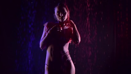 wet sexual slim woman is walking in dark studio with falling water flows, blue and red lamps are lighting on her body
