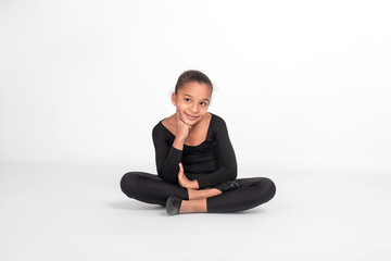 Obraz premium Studio shot of attractive little girl gymnast sitting on the floor with her legs crossed and thinking about something pleasant and holding a finger at her cheek