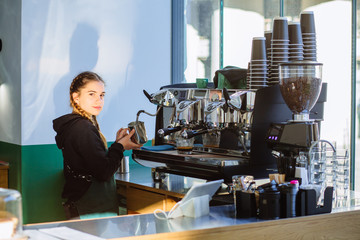 Portrait of hipster young woman working professional barista while preparing coffee beverage for client on modern machine. Positive female waiter with two braids in green apron working intently.