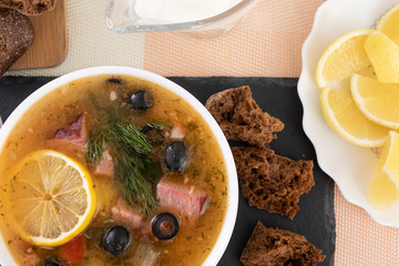 Traditional Russian soup Solyanka cooked with meat, sausages, salted cucumbers on wite bowl. Black bread, lemon, olived and tomatoes on color blue background, homemade food.
