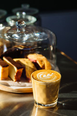 Vertical close up, glass with latte on counter with sweet cakes in glass cake display.