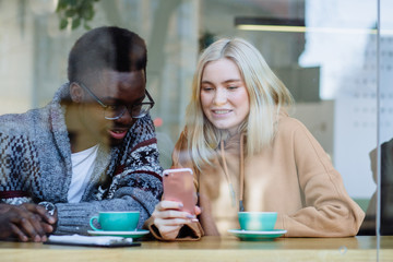 Mixed race couple relax during coffee in cafe. Blond woman and african american man sits together, drinks cappuccino, looks at smartphone near window in winter time. Trough the window.