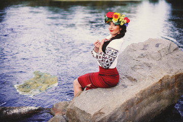 Beautiful woman with dark long hair in national dress red skirt and a white shirt sitting on the stone near river