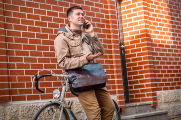 Fototapeta na wymiar Young stylish businessman with his smartphone in hand stay near red wall with sport retro bike in sunny day. 