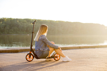 Beautiful sexy blonde woman in denim clothes sits on her scooter on the waterfront street.