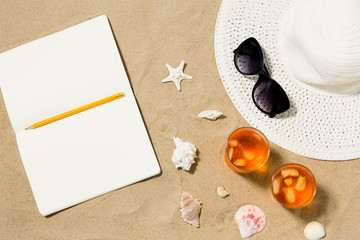 vacation, travel and summer concept - notebook with pencil, two glasses of aperitif cocktails, sun hat and sunglasses on beach sand