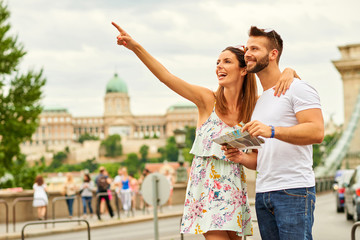 A beautiful young woman is pointing away while her boyfriend is holding a map with the castle of Buda and the Chain Bridge behind them in Budapest, Hungary.
