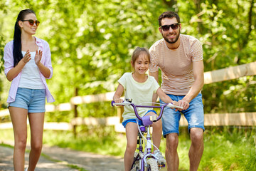 family, parenthood, leisure and people concept - happy mother, father and little daughter learning to ride bicycle in summer park
