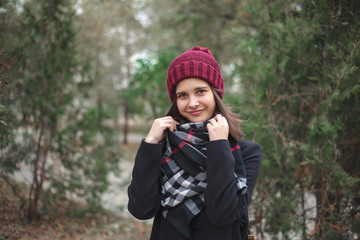 The girl in the red cap straightens the scarf with her hands, looking into the camera and smiling. A portrait of a cute girl.