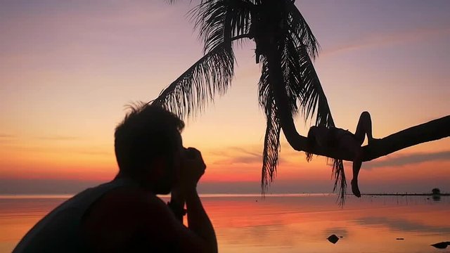 Silhouette of young couple on the beach. Man takes a picture his girlfriend while girl lies on a palm tree during amazing sunset. slwo motion. 1920x1080. People enjoying vacations concept