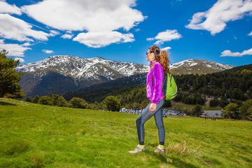 Naklejka premium sporty woman with purple jacket, standing in a green meadow ready to hike, looking at snowy mountain of the horizon, in Cotos (Guadarrama Park, Madrid, Spain)