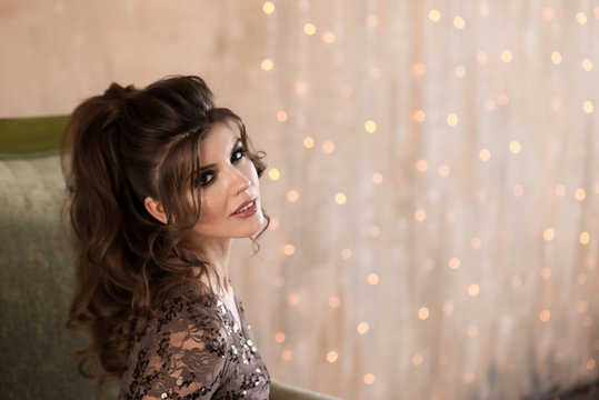 Portrait of a beautiful young girl in a dress with sequins. Hand on face. Festive hairstyle and makeup. Flickering lights on the background.