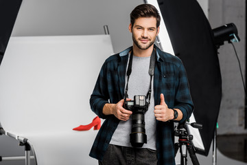 handsome smiling young photographer holding camera and showing thumb up in studio
