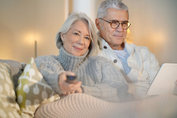  Senior couple at home relaxing on couch watching TV and looking at tablet