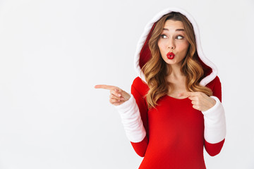Fototapeta premium Image of beautiful woman 20s wearing Christmas red dress pointing fingers at copyspace while standing, isolated over white background