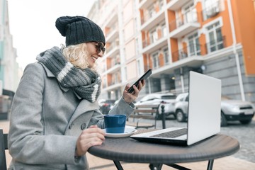 Young woman blogger freelancer in warm clothes hat in outdoor cafe with computer laptop, mobile phone. Urban background