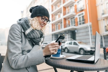 Young woman blogger freelancer in warm clothes hat in outdoor cafe with computer laptop, mobile phone, city street background