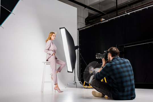 young photographer sitting and photographing attractive stylish female model in studio