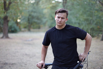 Young handsome caucasian man stands with his bicycle in the park or forest. Thoughful, frowns. White earphones, earring, ring, dark casual wear. Abandoned park background. Outdoors, copy space.