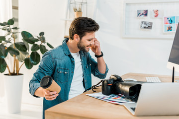 smiling young retoucher holding coffee to go and talking by smartphone at workplace