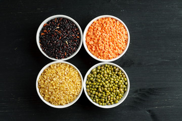 Set of different cereals and legumes in a white bowls on a black table, top view
