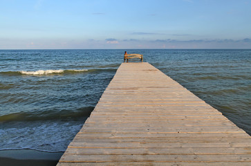 The man sits on the bridge and looks into the sea. A long wooden bridge stretches into the sea