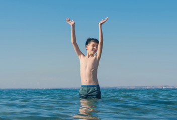 13 years old boy raises his hands standing up in  the sea waves. Concept of family summer vacation