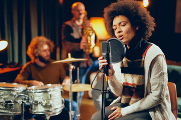 Mixed race woman singing. In background band playing instruments. Home studio interior.