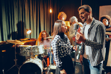 Singers practicing for duet. In background band playing. Home studio interior.