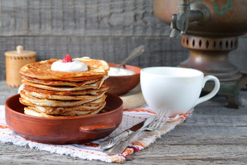 A stack of pancakes in a clay bowl near the old samovar on the gray wooden background.