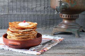 A stack of pancakes in a clay bowl near the old samovar on the gray wooden background.