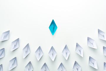 Leadership concept with blue paper ship leading among group of white ships on white background.