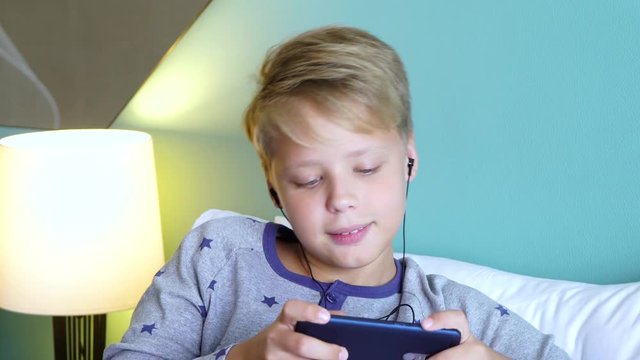 Closeup portrait of cute preteen kid listening to music and singing. Boy laying in bed and holding modern mobile smartphone in hands. Real time 4k video footage.