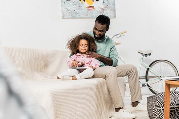 african american father and daughter comfortably sitting on couch and using digital tablet