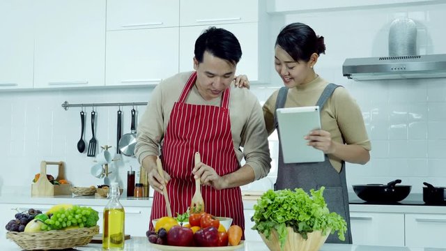 Husband and wife cooking healthy food together at kitchen. Woman assist her husband by using tablet to find technique for cooking salad. People with healthcare concept.