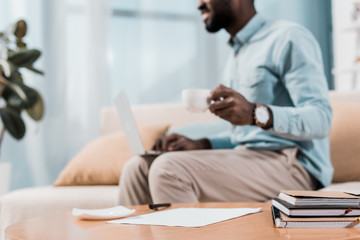 cropped view of african american freelancer sitting on couch with laptop and drinking coffee