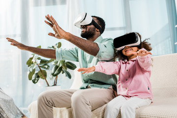 african american father and daughter using virtual reality glasses and smiling in living room