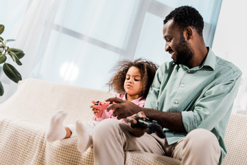 african american dad and daughter sitting on sofa and playing video game in living room