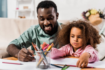 close up view of african american dad helping daughter with drawing in living room