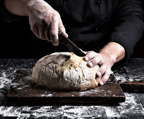 man cuts with a knife a round whole loaf of white wheat flour