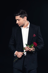 handsome man in suit holding red rose and looking away isolated on black