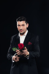 handsome man in suit looking at camera and holding red rose isolated on black