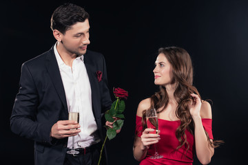 beautiful couple looking at each other, holding red rose and champagne glasses isolated on black