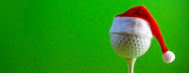 The golf ball mounted on the tee is wearing a Santa Claus souvenir hat. Blank for a postcard for a golfer to celebrate the New Year and Christmas. Bright green background. Copy space.