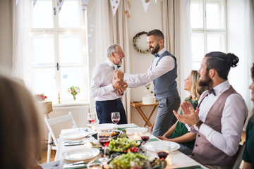 A man giving a bottle of wine to his father on indoor birthday party, a celebration concept.