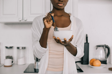 cropped view of african american woman holding spoon and bowl with cornflakes in white kitchen