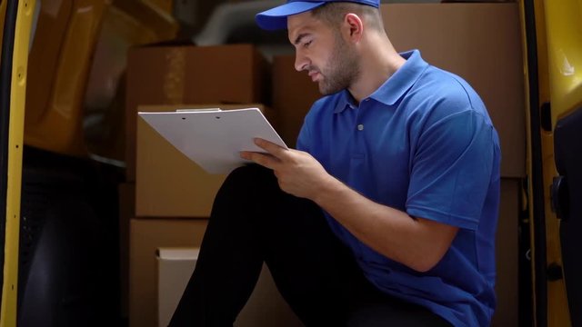 Delivery Man Is Sitting At The Back Of Yellow Truck, Filling Documents