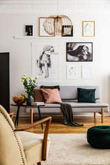 Posters above grey sofa with pillows in living room interior with flowers and armchair. Real photo